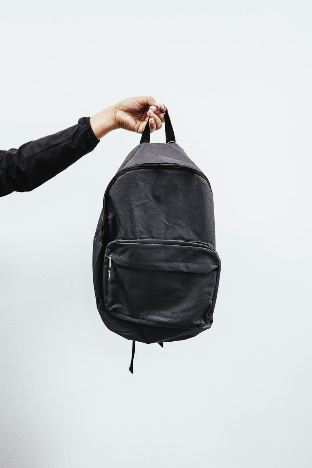 Hand holding a jet-black daypack by its top handle against a white wall