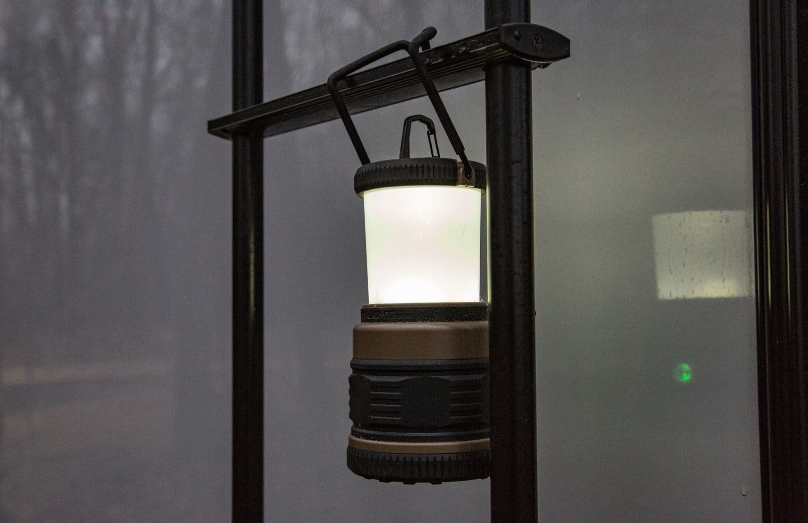 Graphite camp lantern glowing while hanging from a black metal rail by its folding hook