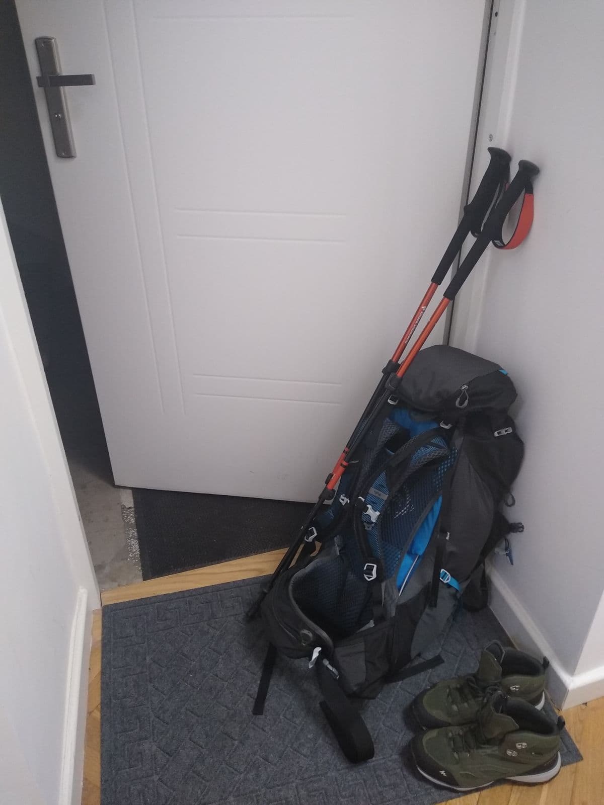Overland 55L Trekking Pack, alternate view: Blue and black trekking pack and two trekking poles leaning in a hallway corner beside hiking shoes