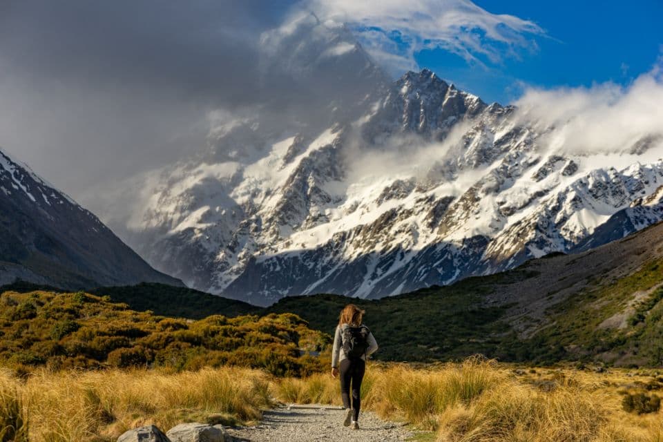 Traverse 32L Pack, alternate view: Hiker with a backpack walking a gravel trail through golden tussock toward snowy peaks