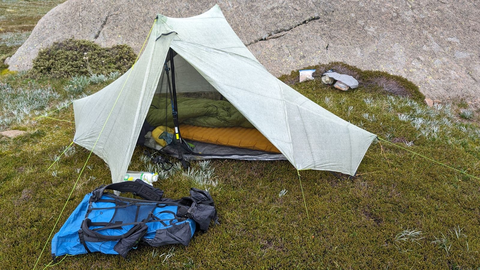 Pale-sage single-pole ultralight shelter pitched on a mossy slope with a blue pack beside it