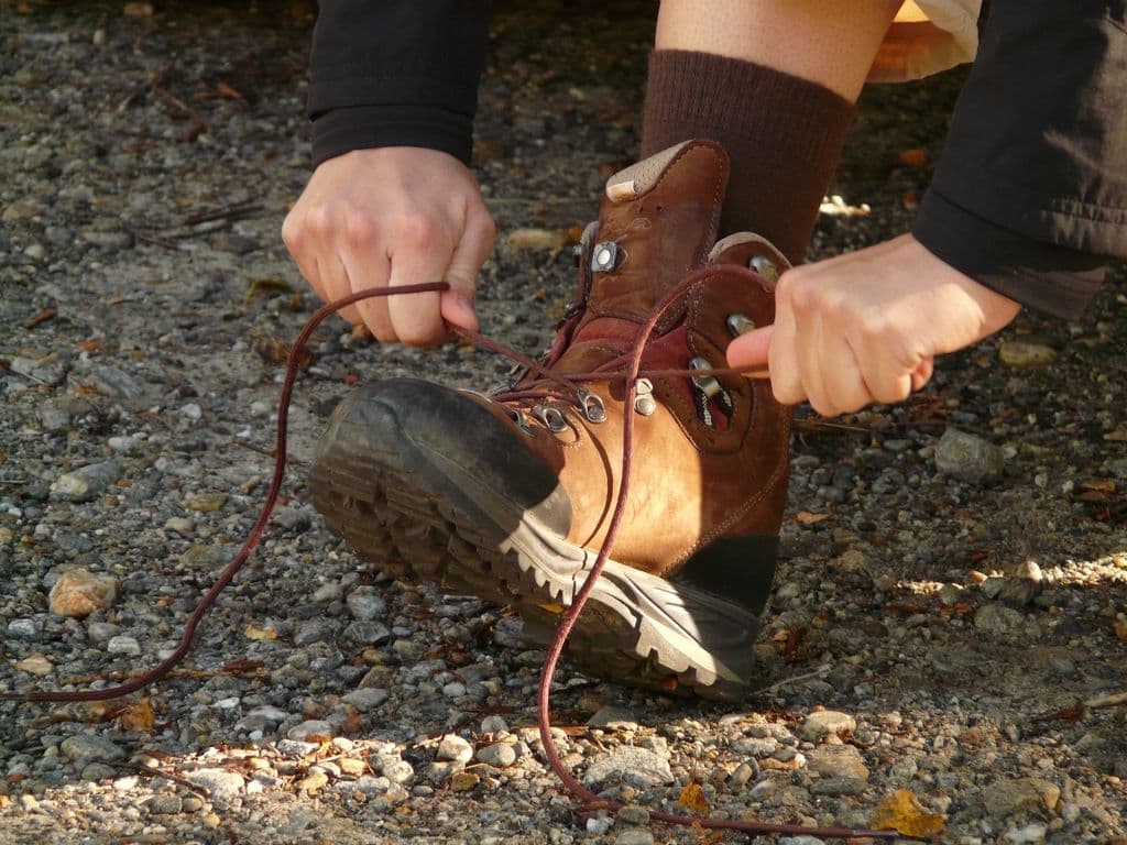 Hands pulling tight the laces of a chestnut leather hiking boot on a gravel trail