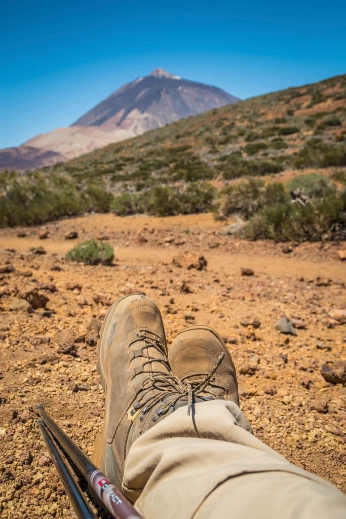 Thumbnail: Desert-tan boots crossed at the ankle on red dirt with a volcano in the distance